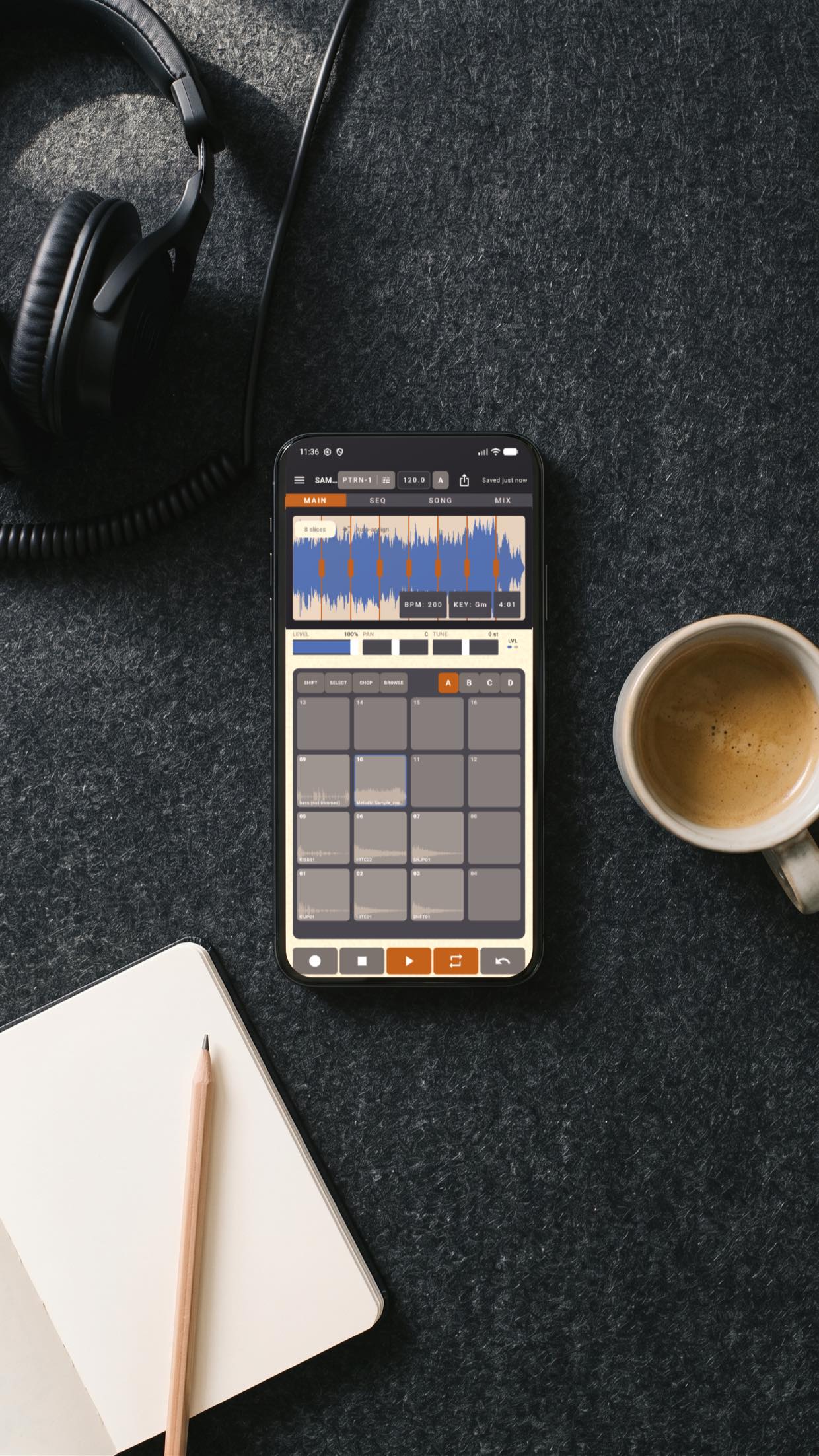 Overhead shot of a phone running SAM PL-1000 on a charcoal felt desk mat: MAIN mode showing a chopped sample waveform at 200 BPM in G minor above the sixteen-pad grid, with record, play and loop buttons along the bottom. Headphones, a notebook and a coffee sit around it.