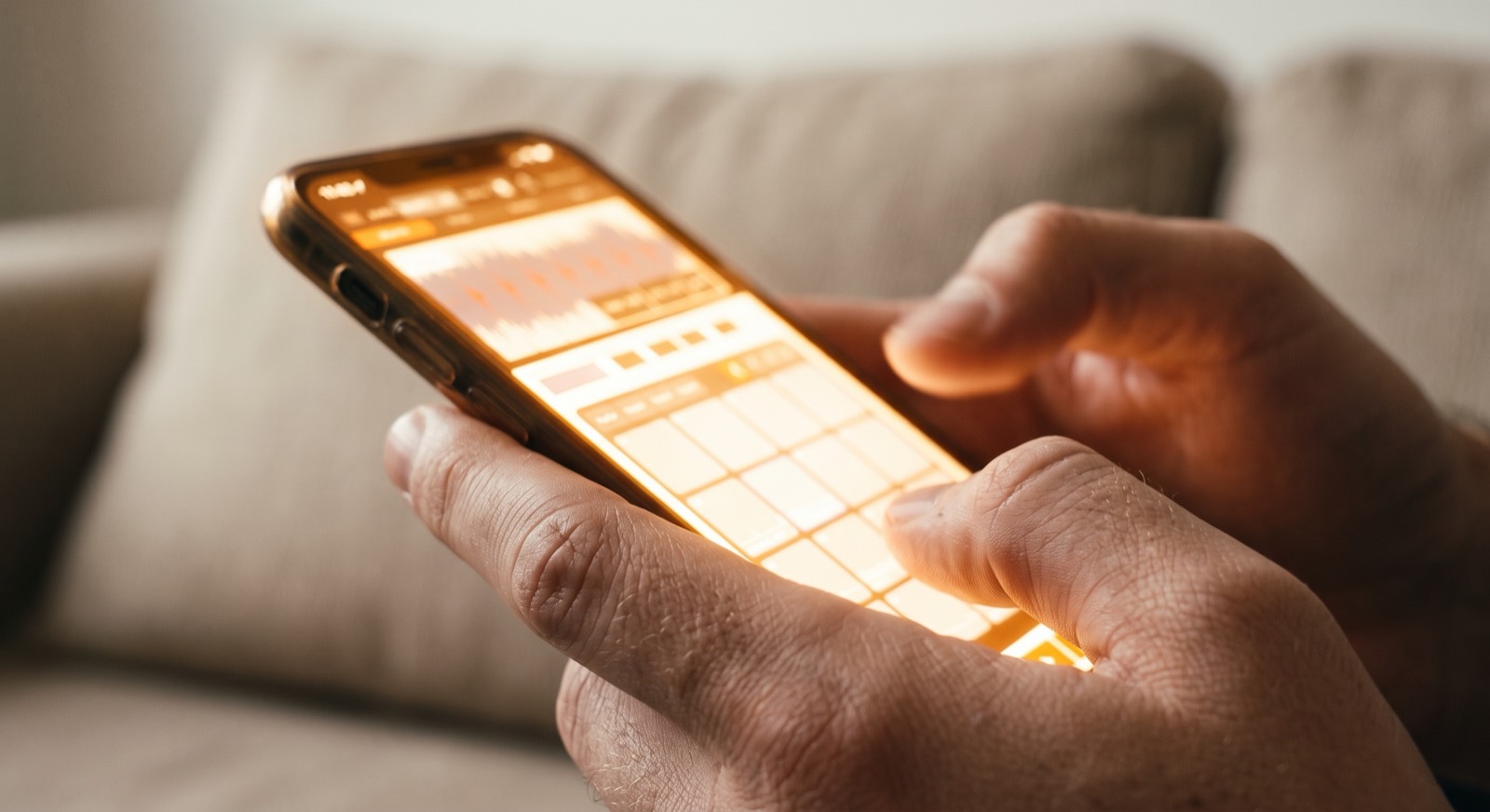 Close-up of two hands holding a phone, thumbs over the pad grid, the screen glow blown out to warm amber across the fingertips.